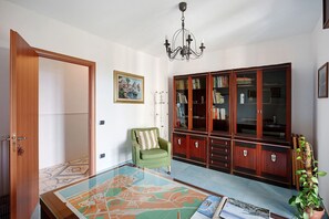 A room with a wooden door, a green chair, a glass coffee table with a map, a bookshelf, and a chandelier.