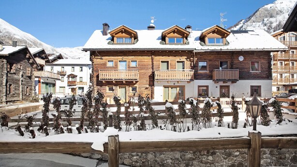 A wooden chalet-style building with balconies, a stone wall, and snow-covered ground. - Mountain Apartment - Rasia Residence Relax (Livigno)