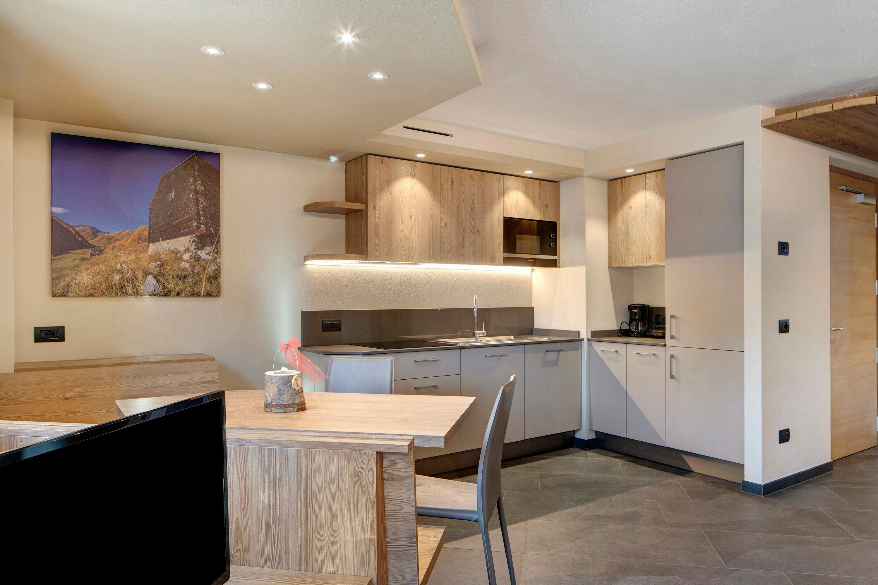 Een moderne keuken met houten kastjes, een eethoek met een tafel en stoelen, en een groot landschap op de muur.