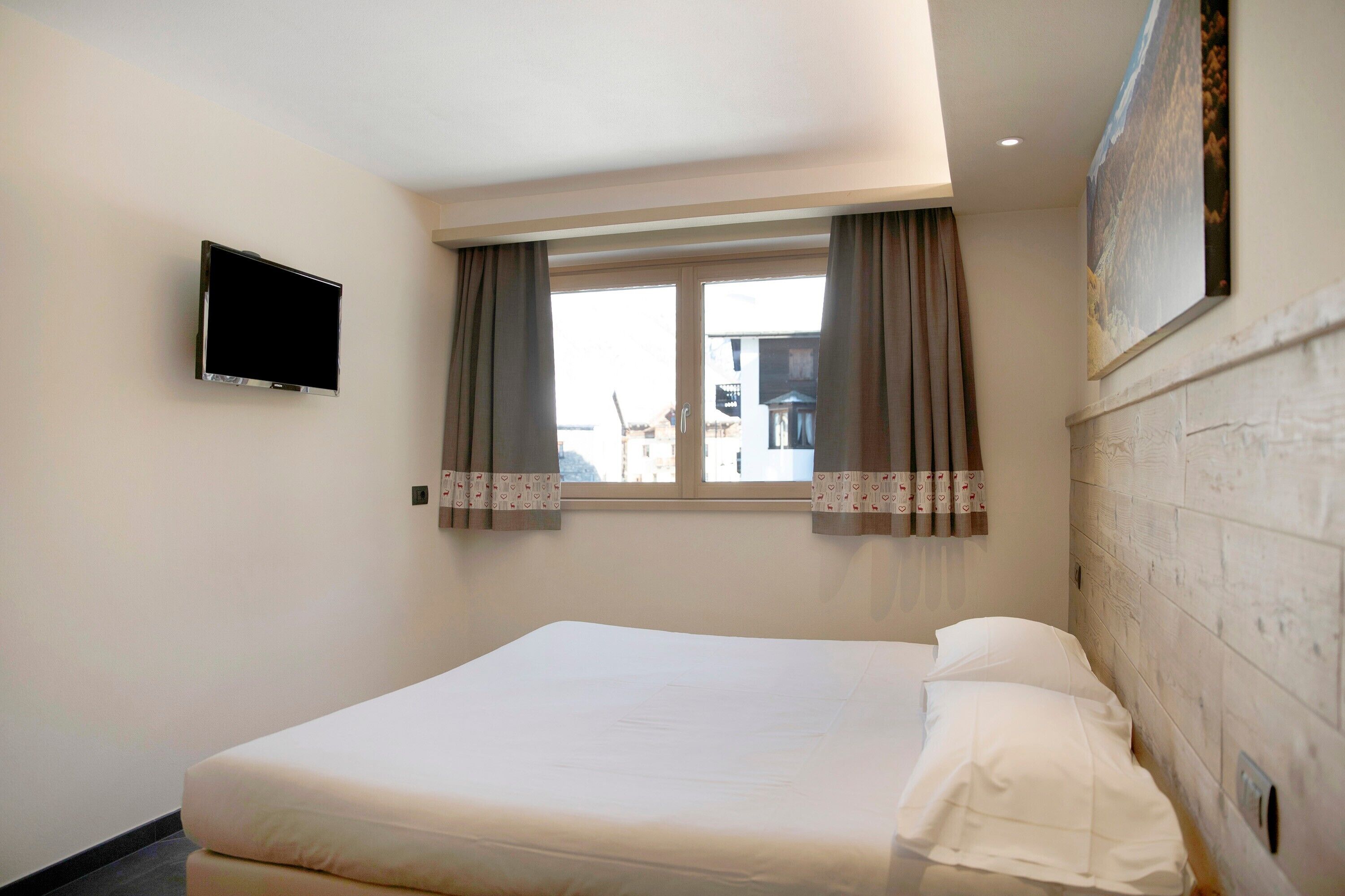 Hotellihuone, jossa on sänky, seinälle asennettu televisio, ikkuna verhoilla ja näkymä ulos.