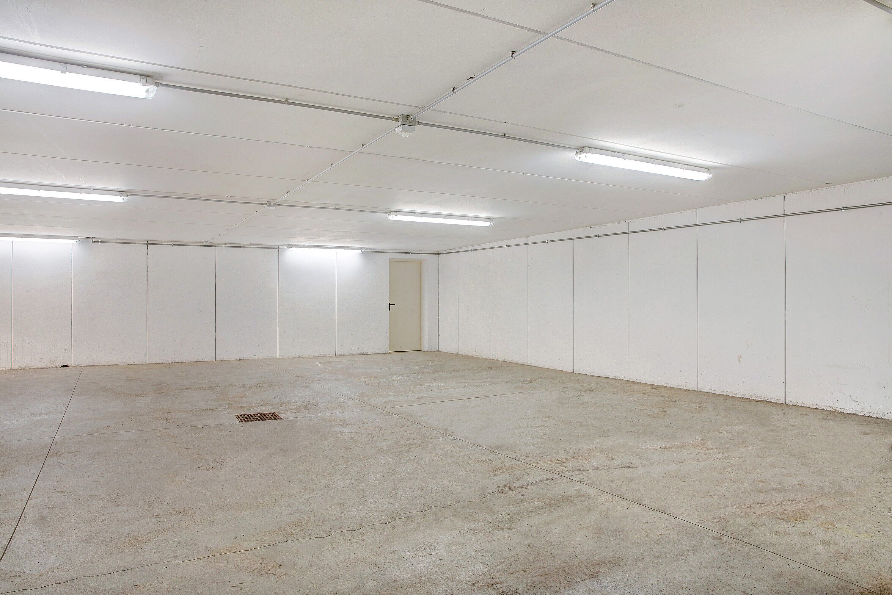 A large, empty room with white walls and a concrete floor, equipped with fluorescent ceiling lights.