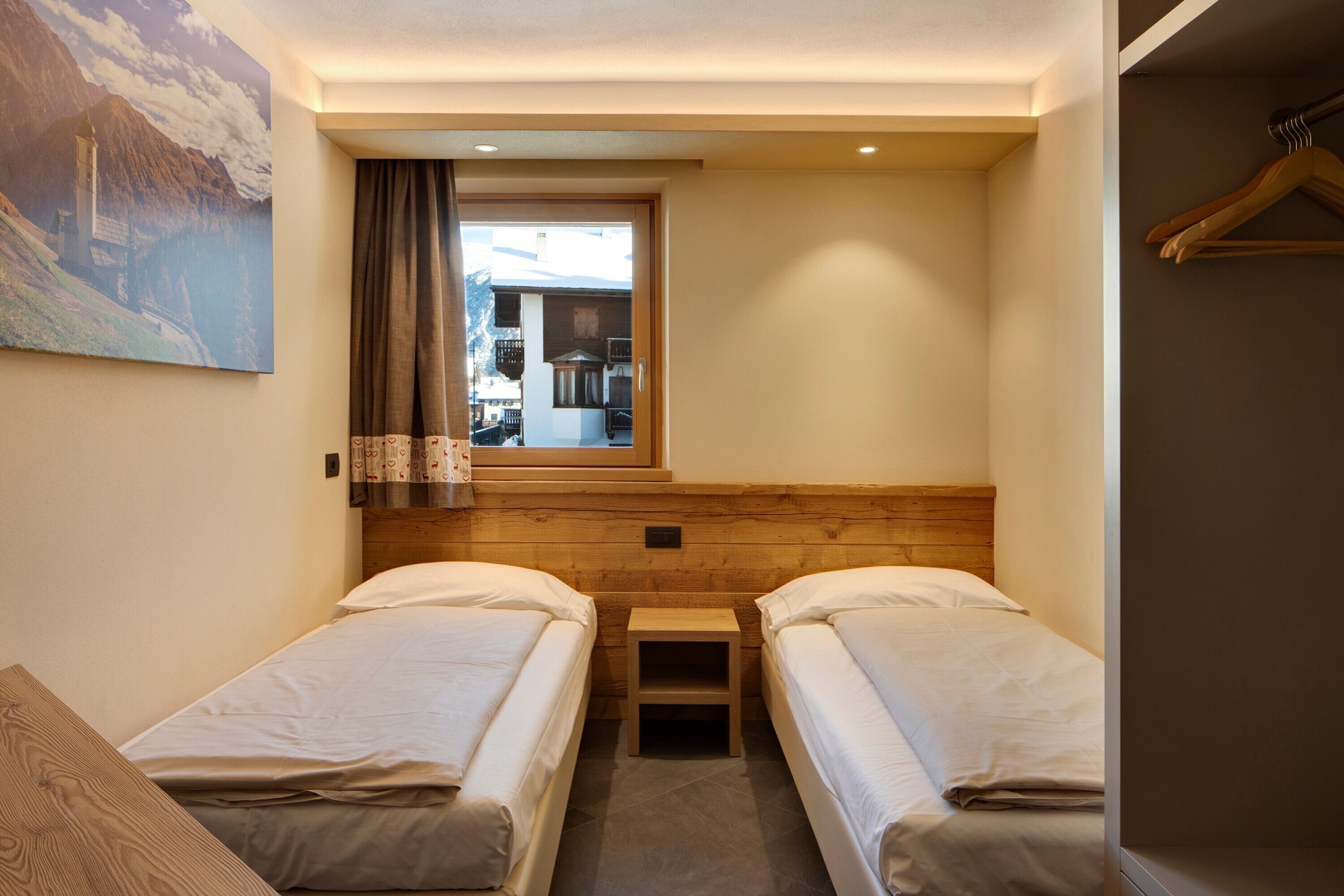 Ein kleines, gemütliches Zimmer mit zwei Einzelbetten, einem Holz-Kopfteil, einem kleinen Tisch und einem Fenster mit Vorhängen.