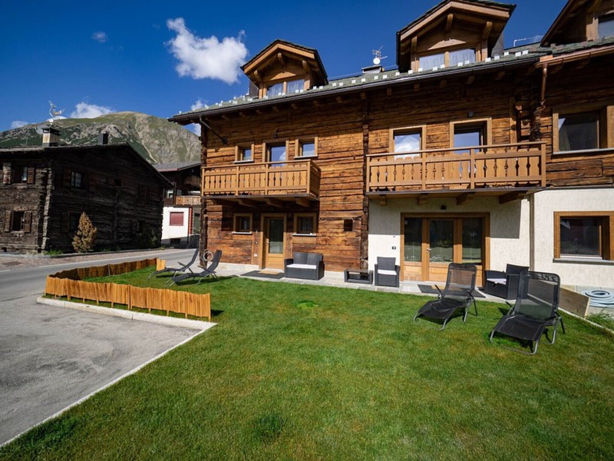 A wooden chalet with a balcony, a lawn with lounge chairs, and a mountain backdrop.