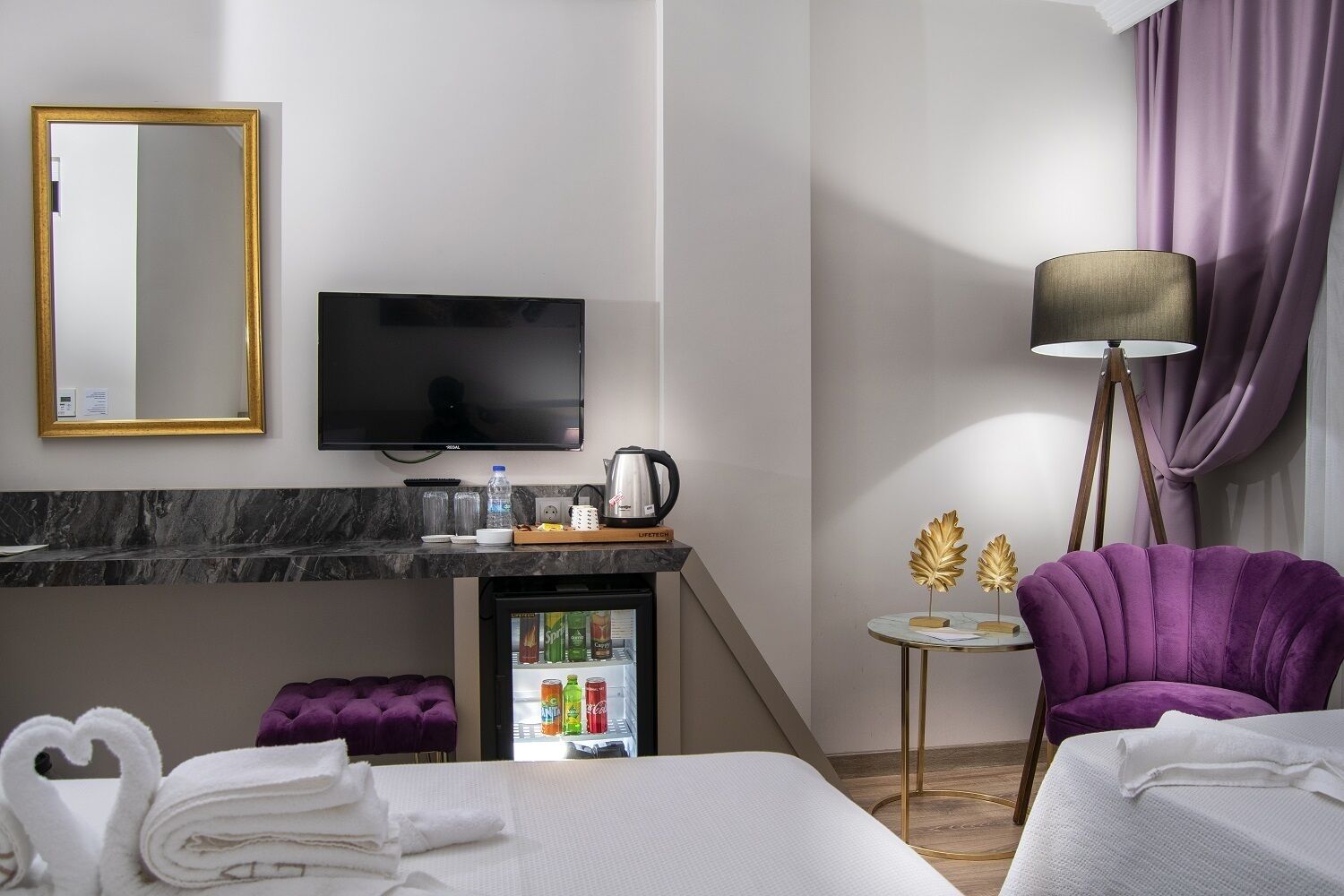 Una habitación de hotel con cama, televisor, minibar, un sillón púrpura y una lámpara.
