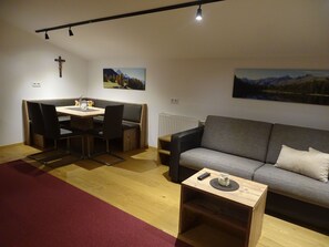 Un salon avec un canapé gris, une table à manger en bois et des chaises. Deux tableaux sont accrochés aux murs : une croix à gauche et un radiateur sous le canapé.