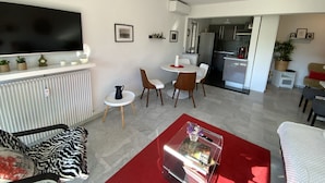 Ein modernes Wohnzimmer mit Flachbildfernseher, eine Essecke mit weißem Tisch und Stühlen, eine Küche mit Kühlschrank und Herd sowie eine rote Akzentwand.