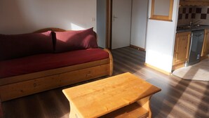 Ein kompakter Wohnbereich mit einer Couch, einem kleinen Holztisch, einer Küchenzeile und einer Tür zu einem weiteren Raum.