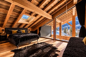 Quarto com teto inclinado, uma cama, uma janela grande e vista para as montanhas.