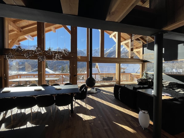 A spacious room with large windows offering mountain views, wooden beams, and a dining area with chairs and a table.
