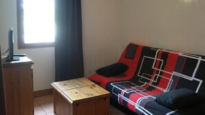 Ein kleiner Raum mit einem Holztisch, einer Couch mit gemustertem Bezug, einem Fenster mit Vorhängen und einer Wandleuchte.