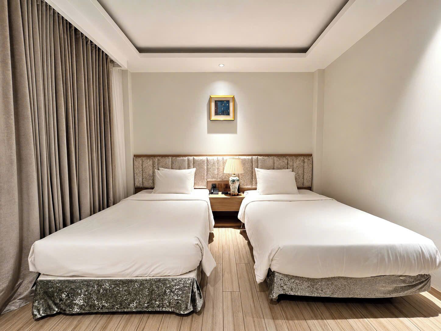 A hotel room with two beds, a nightstand with a lamp, a framed picture on the wall, and a window with curtains.