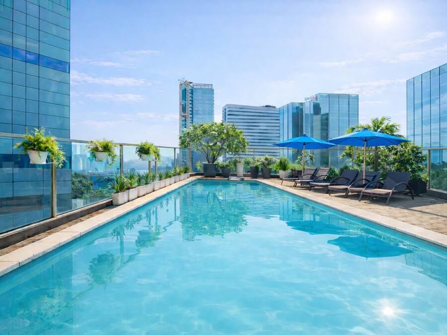 Una piscina en la azotea con agua azul cristalina, rodeada de sillones y sombrillas, y edificios modernos al fondo.