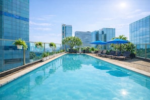 A rooftop pool with clear blue water, surrounded by lounge chairs and umbrellas, with modern buildings in the background.