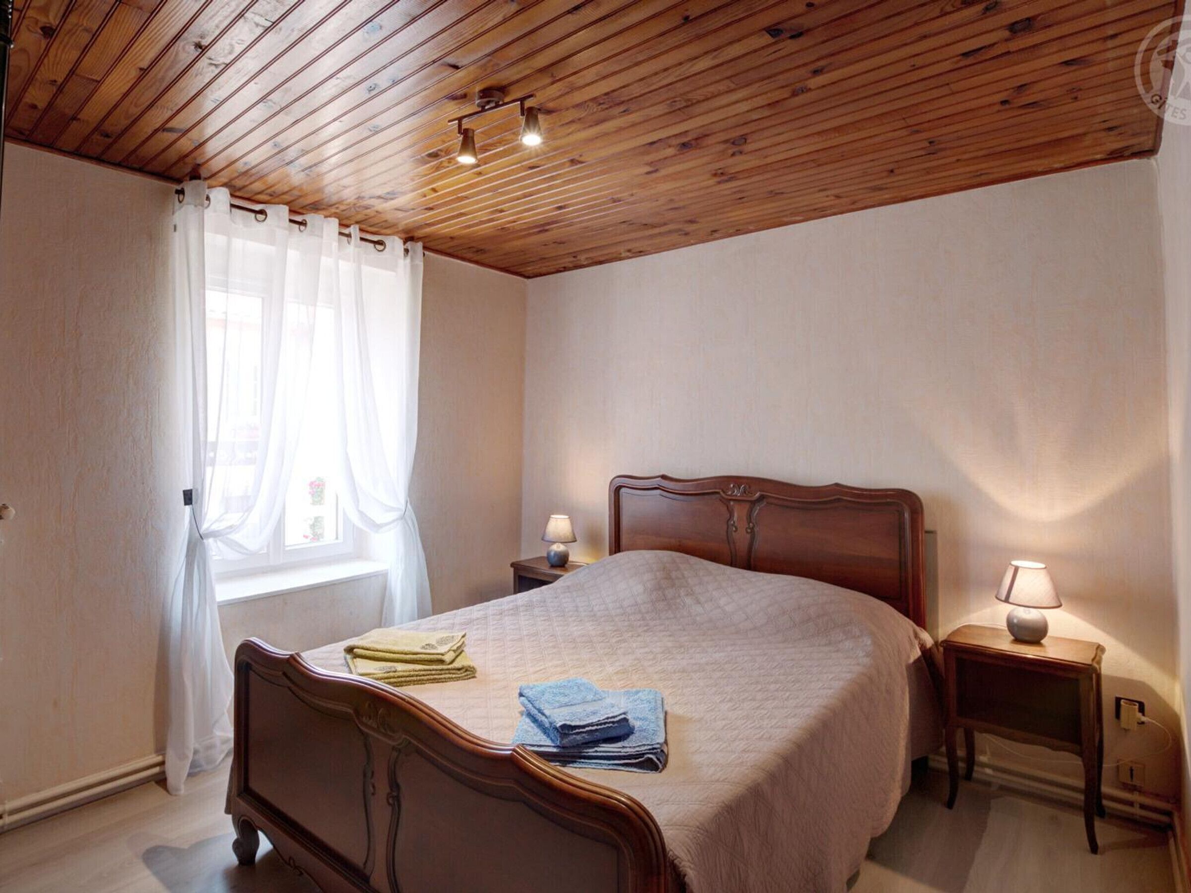 A bedroom with a wooden ceiling, a bed with a quilt, two bedside tables with lamps, and a window with sheer curtains.