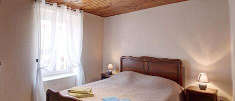 A bedroom with a wooden ceiling, a bed with a quilt, two bedside tables with lamps, and a window with sheer curtains.