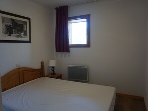 A bedroom with a bed, a nightstand with a lamp, a radiator, a window with curtains, and a framed picture on the wall.