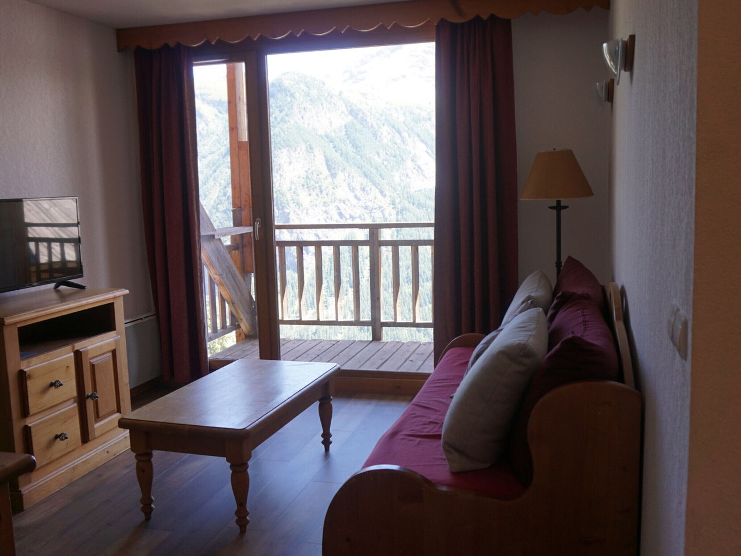 Een kamer met een houten tafel, een bank met kussens, een televisie op een houten kast en een balkon met uitzicht op de bergen.