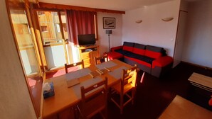 A living room with a red and black sofa, a wooden dining table with chairs, a television on a wooden stand, and a window with curtains. - Bright apartment with balcony, parking, WIFI, near slopes (Orcières Merlette)