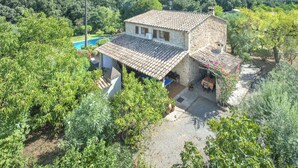 A stone house with a tiled roof, surrounded by lush greenery and a swimming pool.