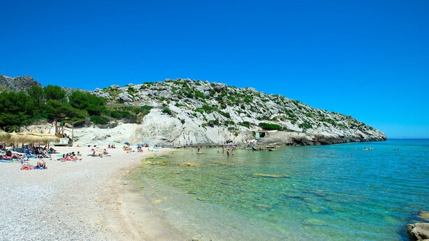 A beach with clear water, people sunbathing, and a rocky hill in the background.