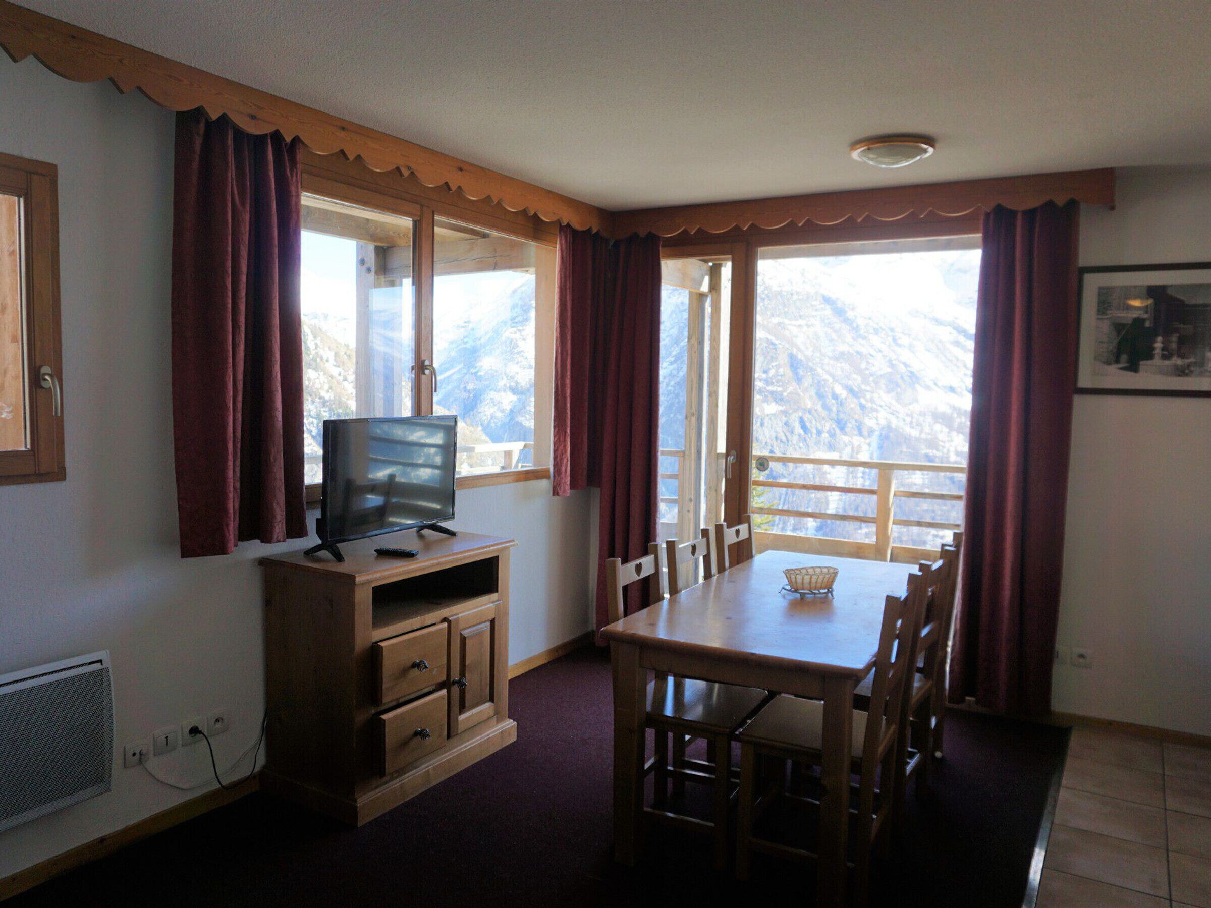 A room with a wooden TV stand, a dining table with chairs, and a view of mountains through large windows.