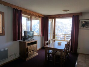 A room with a wooden TV stand, a dining table with chairs, and a view of mountains through large windows.