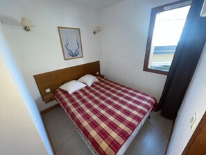Ein Einzelbett mit karierter Bettdecke in einem Raum mit weißen Wänden, einem Holz-Kopfteil, zwei Wandlampen und einem Bild von Geweihen im Bilderrahmen.