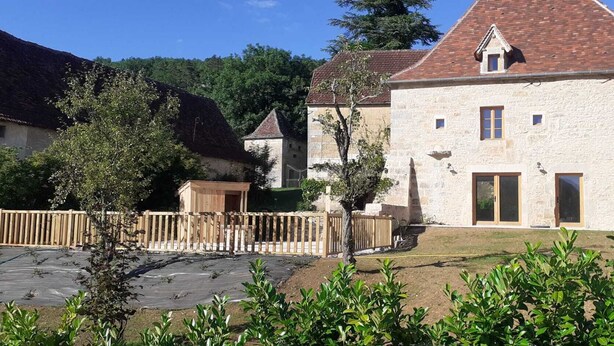 Un edificio de piedra con techo de tejas rojas, cerca de madera y una pequeña construcción de madera.