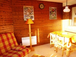 A cozy cabin interior with a wooden wall, a plaid sofa, a dining table with chairs, and a lamp.