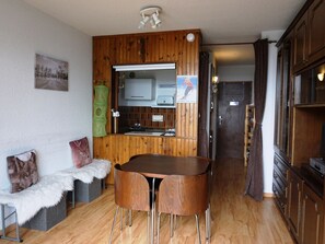 A compact kitchen with wooden cabinets, a dining table with two chairs, a small sink, and a refrigerator.