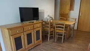 A wooden dining table with chairs, a television on a wooden cabinet, and a kitchen area with cabinets and a window. - 2-room flat, 4 pax, pool, WiFi, covered parking (Orcières Merlette)