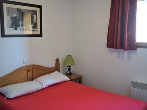 A single bed with a wooden headboard, a green bedside lamp, a red bedspread, a framed picture on the wall, and a window with red curtains. - 3-Room Apartment for 6 in Quality Residence, Close to Shops and Slopes, Free WiFi, Pets Allowed (Orcières Merlette)