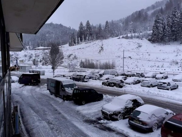 A snowy parking lot with several vehicles, including a truck and cars, parked on a road with a mountainous backdrop.