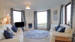 A living room with two white sofas, a round blue rug, a flat-screen TV, a fireplace, and large windows with plaid curtains. - Aurora View (Forres)