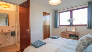 A bedroom with a bed, a bedside table with a mirror, a window with curtains, and a bathroom with a sink and shower. - Aurora View (Forres)