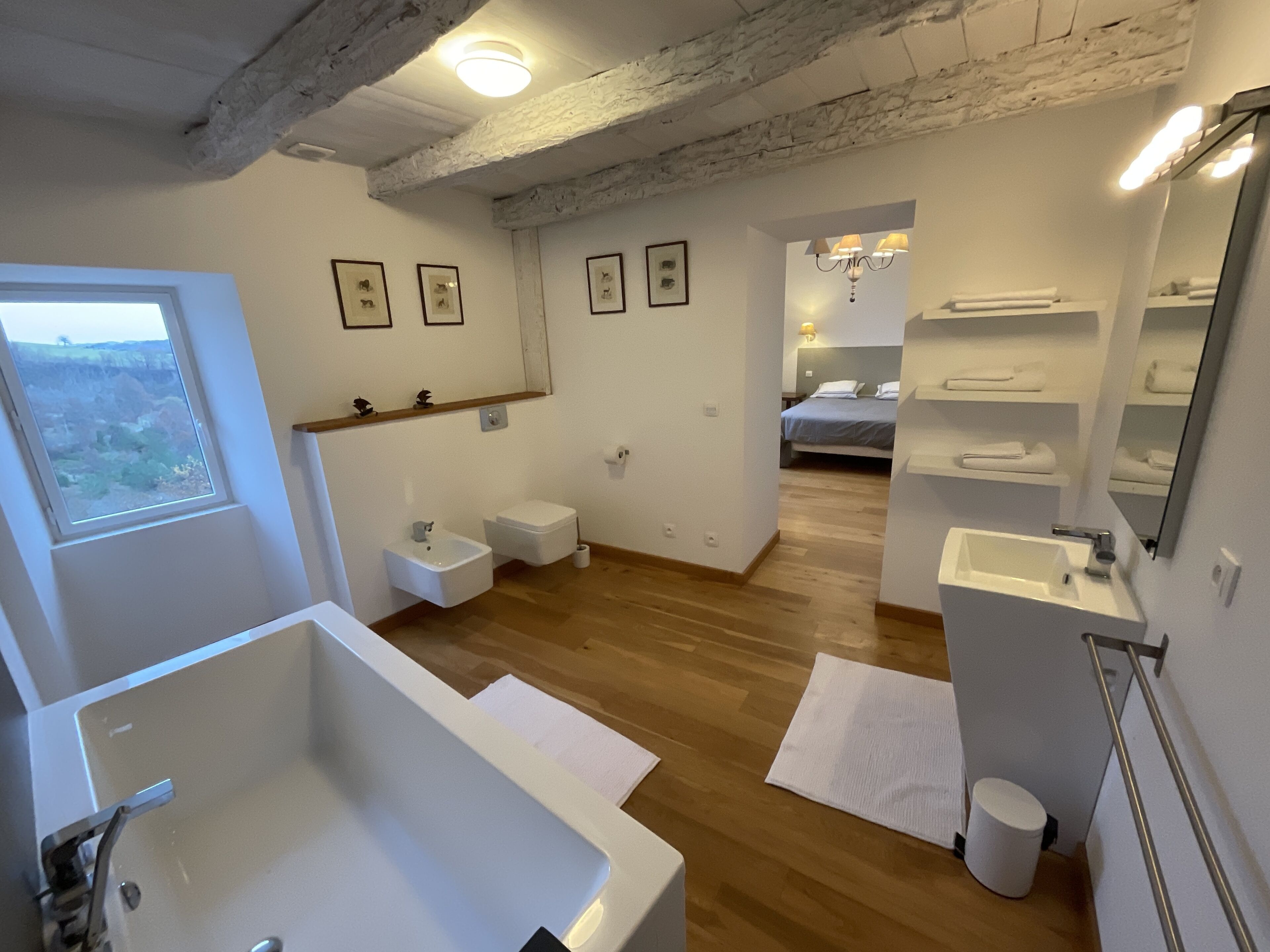 A modern bathroom with a large bathtub, a small sink, and a toilet. There are framed pictures on the wall, a window with a view, and a shower area.