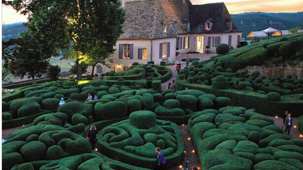 A maze garden with pathways, a building with a stone roof, and people walking. - LE CLOS DE PEYRUZEL (Daglan)