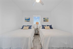A room with two beds, a ceiling fan, a window with blinds, and two wall-mounted paintings.