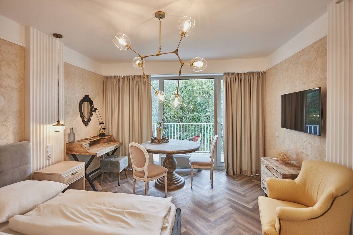 Hotelkamer met een bed, een bureau met stoel, een eettafel met stoelen, een gele fauteuil, een kroonluchter, een aan de muur bevestigde televisie en een balkon met gordijnen.