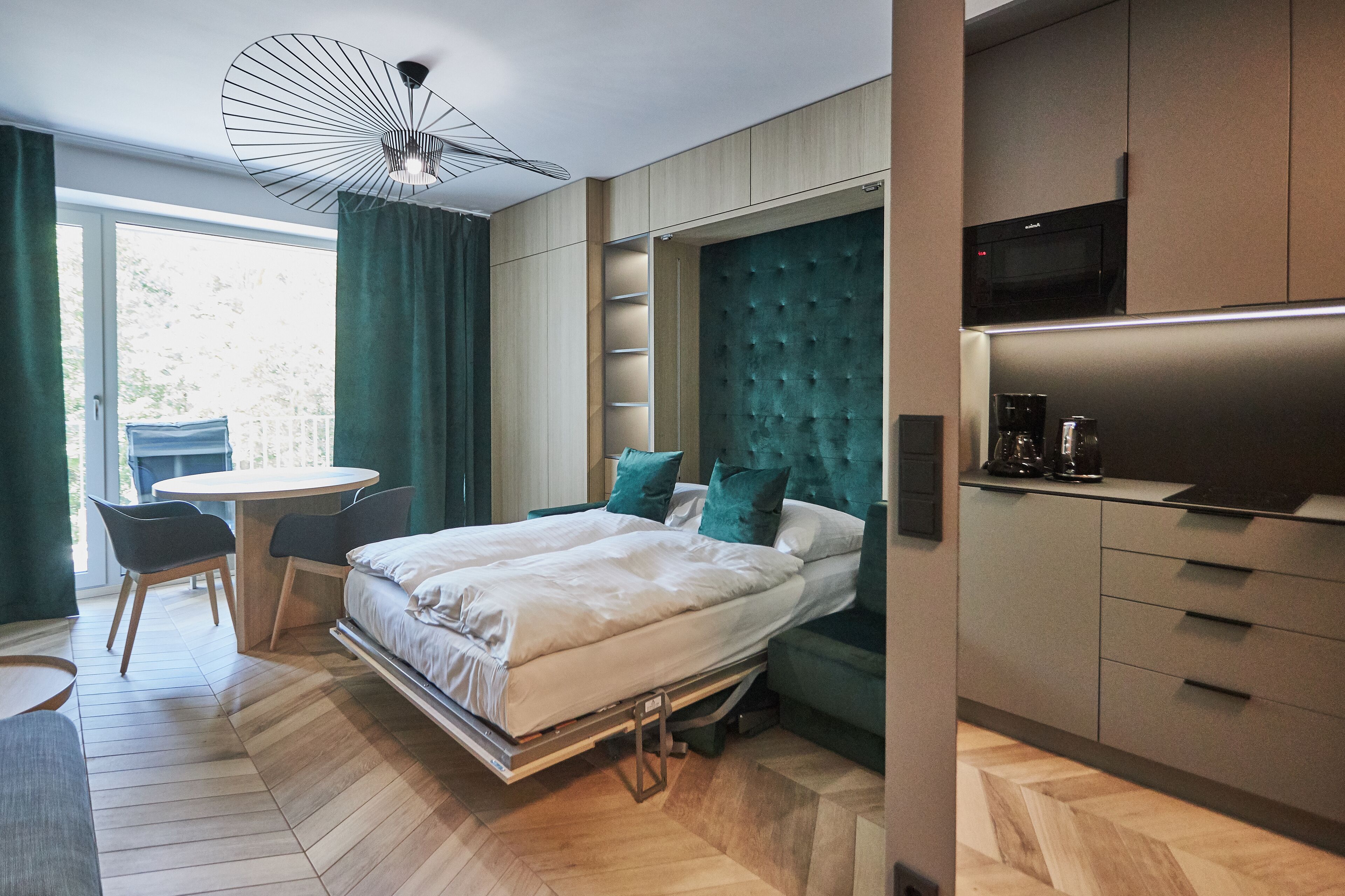 A modern bedroom with a bed, bedside tables, a desk, and a chair. There is a built-in wardrobe and a kitchenette with a microwave.