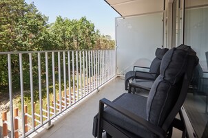 Un balcón con barandilla blanca, dos sillas grises y un ventanal de vidrio que da a una habitación.