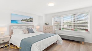 Une chambre à coucher comprenant un lit, des tables de chevet, une commode, un banc et une fenêtre avec des stores.