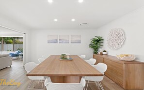 A modern dining area with a wooden table and white chairs, a wooden cabinet, and a decorative wall piece. - Waves on Whitesands - absolutely EVERYTHING for your beach holiday (Fingal Bay)