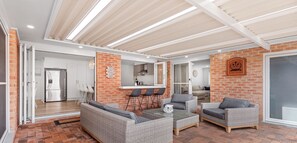 A spacious living area with wicker furniture, a kitchen with a brick wall, and a sliding glass door leading to another room. - Waves on Whitesands - absolutely EVERYTHING for your beach holiday (Fingal Bay)