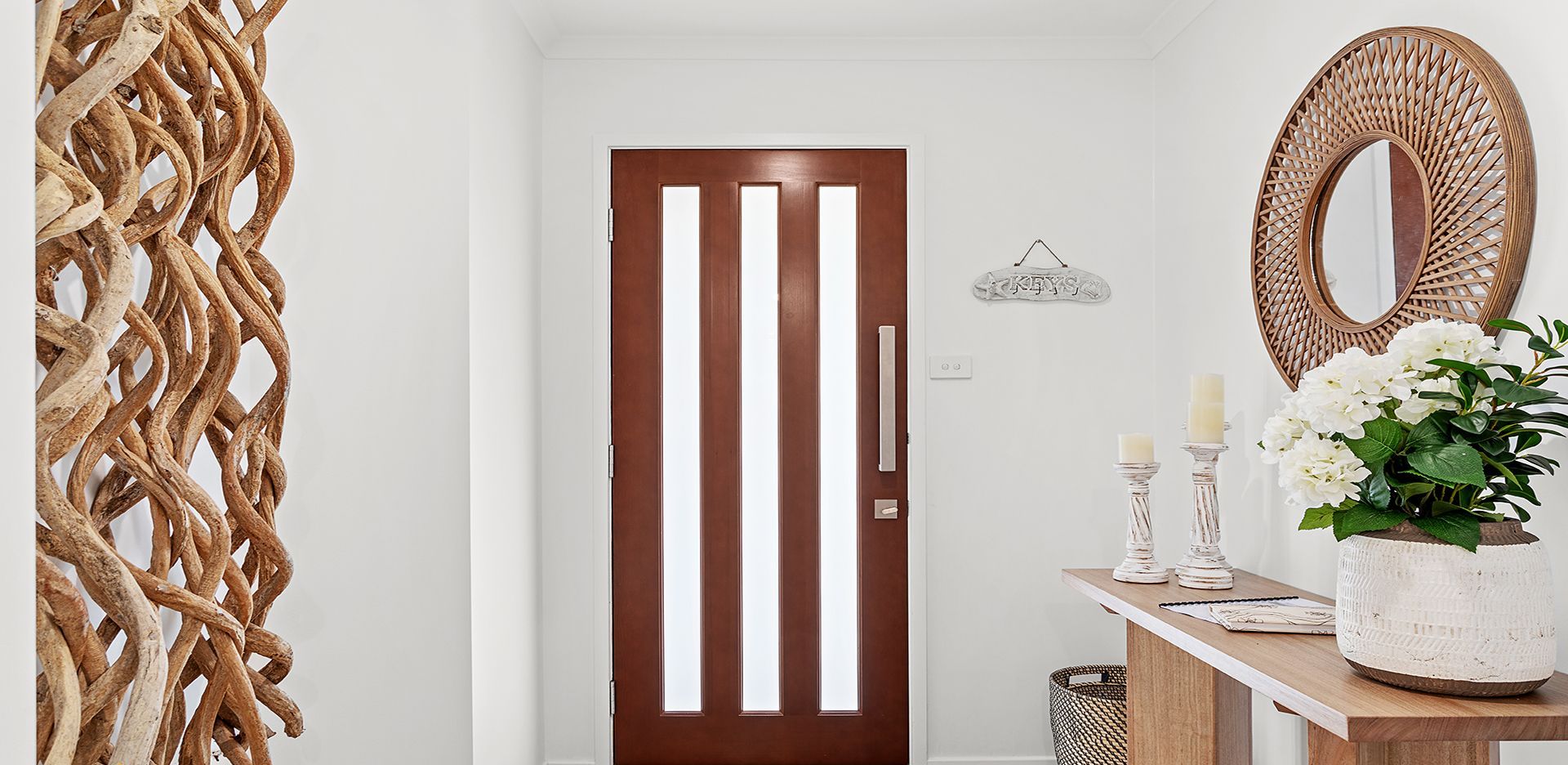 A hallway with a wooden door, a woven wall decoration, a mirror, and a small table with candles and flowers.