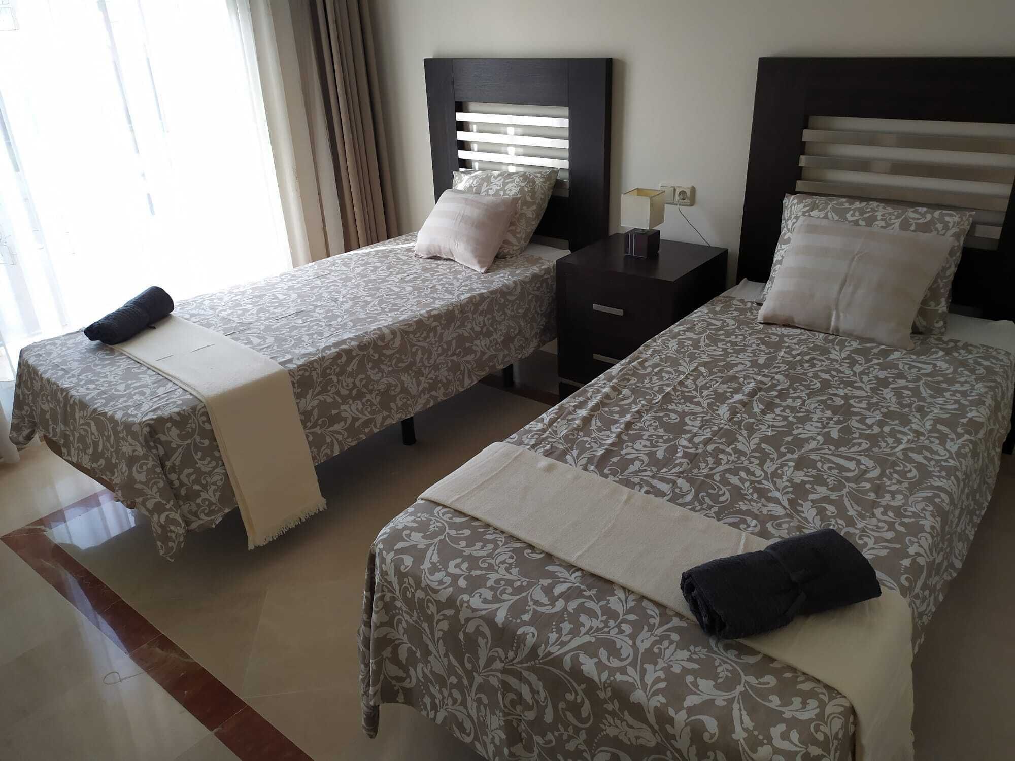 A hotel room with two single beds, each with a bedside table and lamp.