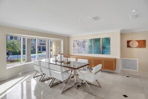 A modern dining area with a glass table and white chairs, large windows with a view of a residential area, and abstract wall art. - Palms by the Sea Villa 🌊 Waterfront | Heated Pool | Near Beach 🏖️ (Fort Lauderdale)