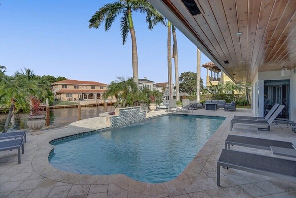 A swimming pool with lounge chairs, palm trees, and a covered area with a wooden ceiling.