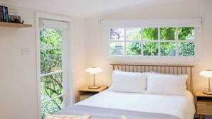 A bedroom with a bed, two bedside tables with lamps, a window with a view of greenery, and a small wooden table with chairs.
