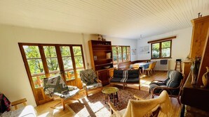Amplia sala de estar con una gran estantería de madera, diversas opciones de asientos y grandes ventanas que permiten la entrada de luz natural.